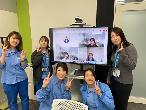 オンライン会議をしている大きなディスプレイの周りでピースサインをしている5名の女性。ディスプレイには4名の女性が写っている